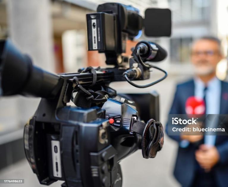 Camera in focus, blurred journalist, dynamic reporting atmosphere.