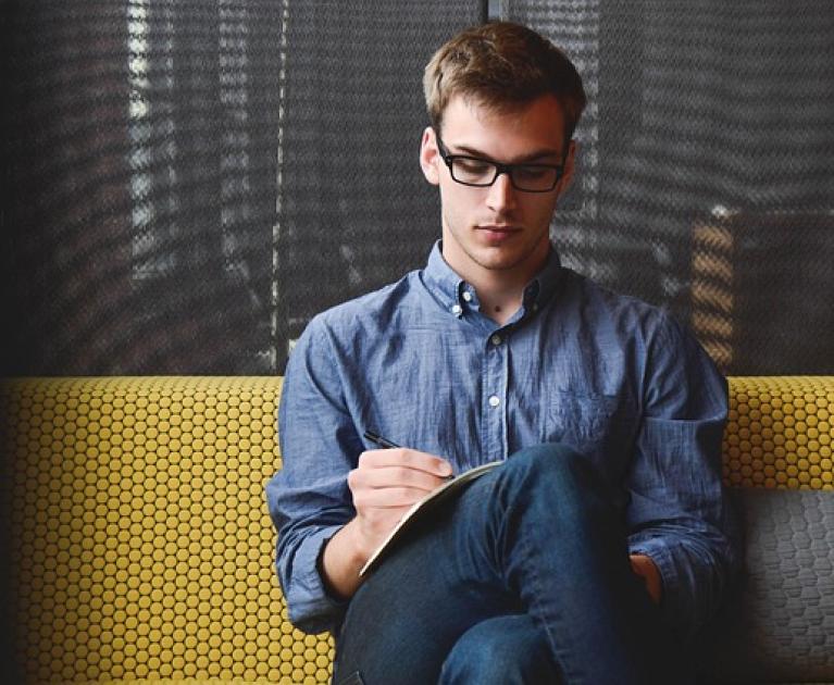 Thoughtful young man writing, sitting on a yellow sofa, calm atmosphere.
