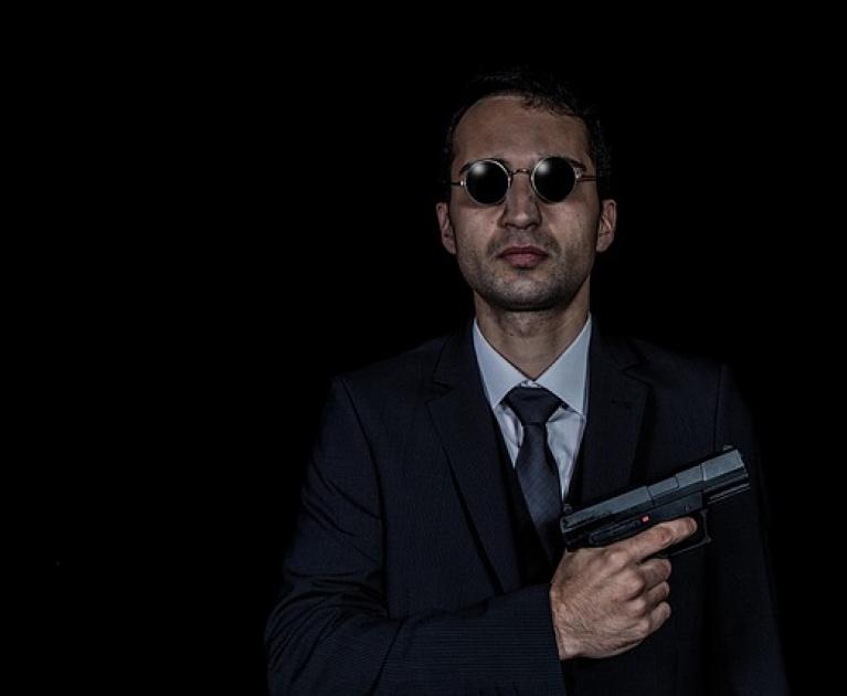 Man in dark suit with glasses, holding a gun, intense and mysterious atmosphere.